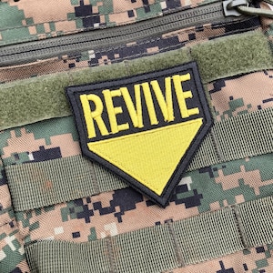 以下が含まれることがあります： 白文字で「REVIVE」と書かれた黒と黄色のパッチ。パッチは迷彩柄の生地に付けられています