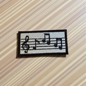 Pode incluir: Um patch retangular com notas musicais e uma clave de sol. O design preto sobre um fundo bege claro, com uma borda preta. Ideal para roupas ou acessórios.