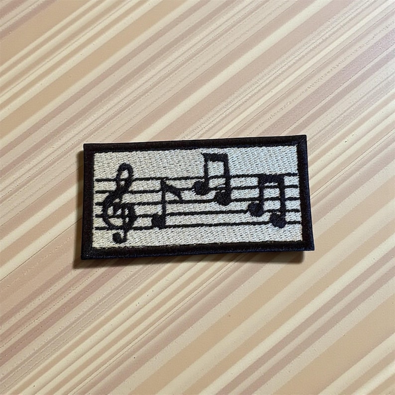 May include: A rectangular patch featuring musical notes and a treble clef. The design is in black against a light beige background with black border. The patch is likely for clothing or accessories.