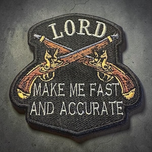 May include: A shield-shaped embroidered patch with a black background featuring two crossed brown muskets. The text LORD is embroidered in white above the muskets, and the text MAKE ME FAST AND ACCURATE is embroidered in white below the muskets.