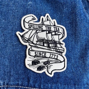 May include: A white embroidered patch with a black illustration of a tall ship and the text "Spilling the Tea Since 1773". The patch is sewn onto a blue denim jacket.