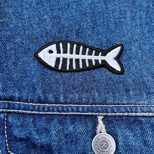 Può includere: Primo piano di una giacca di jeans con una toppa a forma di scheletro di pesce. La toppa è bianca e nera e ha la forma di un pesce. È visibile un bottone argentato con la scritta "TODAYS NEWS".