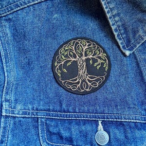 May include: A circular embroidered patch depicting a tree of life, with green leaves and a brown trunk and roots. The patch is attached to a blue denim jacket, placed on the chest.