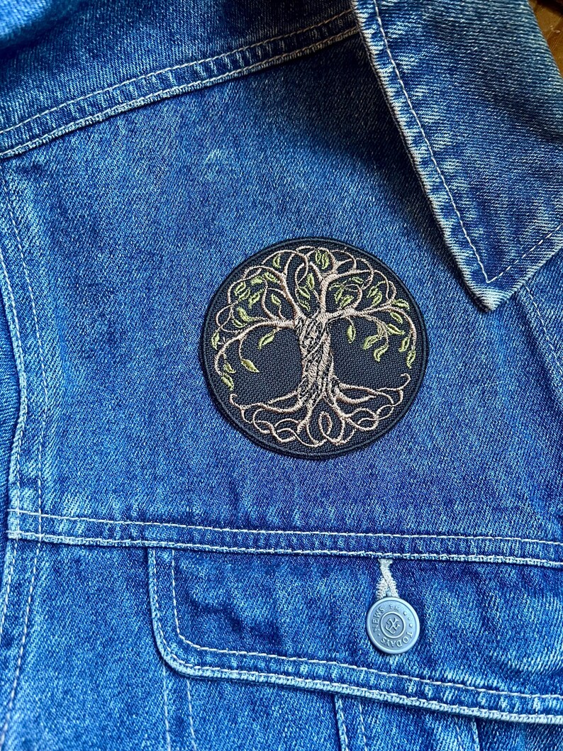 May include: A circular embroidered patch depicting a tree of life, with green leaves and a brown trunk and roots. The patch is attached to a blue denim jacket, placed on the chest.