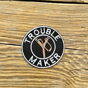 May include: A circular black patch with the words "TROUBLE MAKER" in white around the edge. A tan slingshot design is in the centre. The patch is displayed on a wooden surface.