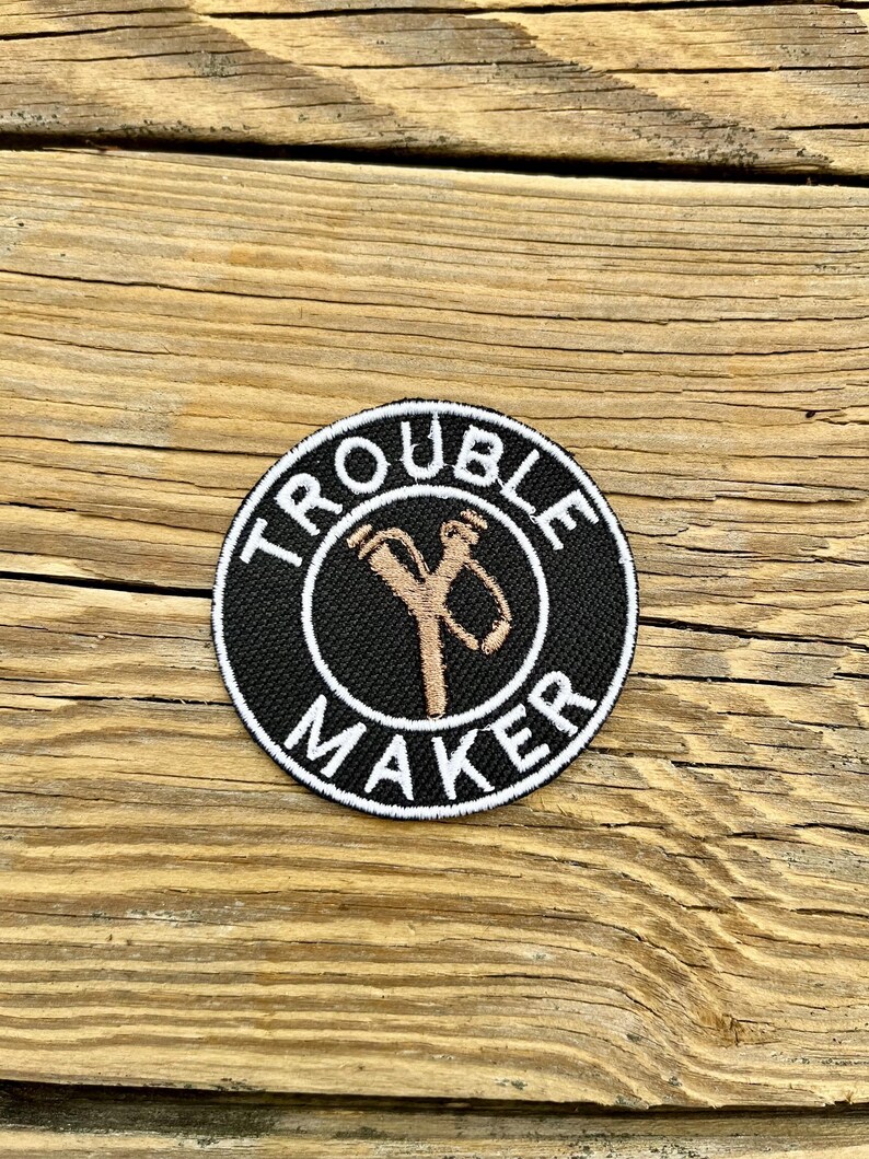 May include: A circular black patch with the words "TROUBLE MAKER" in white around the edge. A tan slingshot design is in the centre. The patch is displayed on a wooden surface.