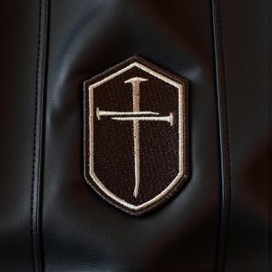 May include: A black leather item featuring a brown and beige embroidered patch. The patch is shield-shaped and displays a cross design with a vertical line and a horizontal line, with a beige border.