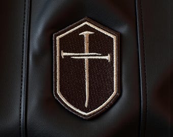 Puede incluir: Un artículo de cuero negro con un parche bordado marrón y beige. El parche tiene forma de escudo y muestra un diseño de cruz con una línea vertical y una horizontal, con un borde beige.