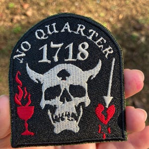 May include: A black embroidered patch featuring a white skull with horns and the text "No Quarter 1718". Below the skull is a red heart with an arrow through it. There are red flames on the left side of the skull and a red goblet.