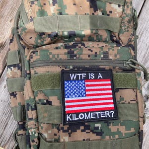 Pode incluir: Uma mochila t&aacute;tica camuflada com um patch que diz "WTF IS A KILOMETER?" e apresenta uma pequena bandeira americana.