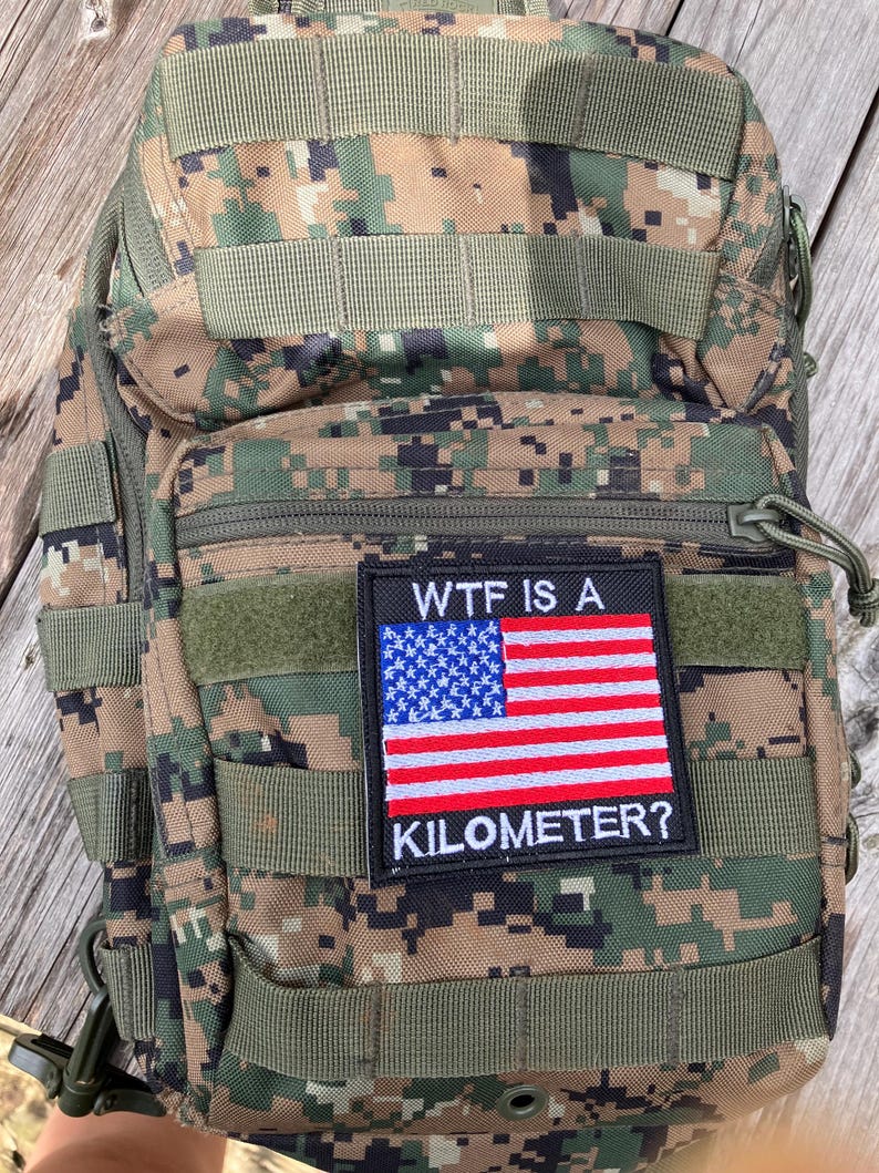 Pode incluir: Uma mochila t&aacute;tica camuflada com um patch que diz "WTF IS A KILOMETER?" e apresenta uma pequena bandeira americana.