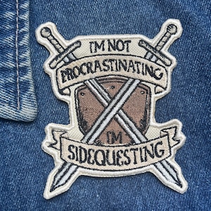 May include: A beige embroidered patch with two crossed swords and a shield. The patch has the text "I'm not procrastinating I'm sidequesting" on a banner.
