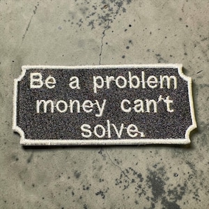 Puede incluir: Un parche rectangular con fondo gris oscuro y texto bordado en blanco que dice "Be a problem money can't solve". El parche tiene un borde blanco y un borde decorativo.