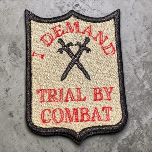Pode incluir: Patch bordado em forma de escudo com as palavras "I DEMAND TRIAL BY COMBAT" em vermelho. Duas espadas cruzadas no centro. O patch tem um fundo bege e uma borda preta.