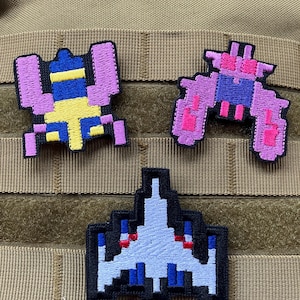 Könnte beinhalten: Drei gestickte Patches mit pixelartigen Videospielfiguren. Der erste Patch ist ein rosa und gelbes Raumschiff, der zweite ist ein lila und rosa Außerirdischer und der dritte ist ein weißes Raumschiff mit roten und blauen Akzenten.