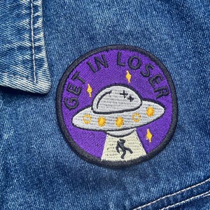Może przedstawiać: Naszywka haftowana w kolorze fioletowym i czarnym z białym UFO i tekstem "Gettin' Loser" w kółku.