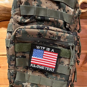 Pode incluir: Uma mochila t&aacute;tica de camuflagem com um patch que diz "WTF IS A KILOMETER?" e apresenta uma pequena bandeira americana.