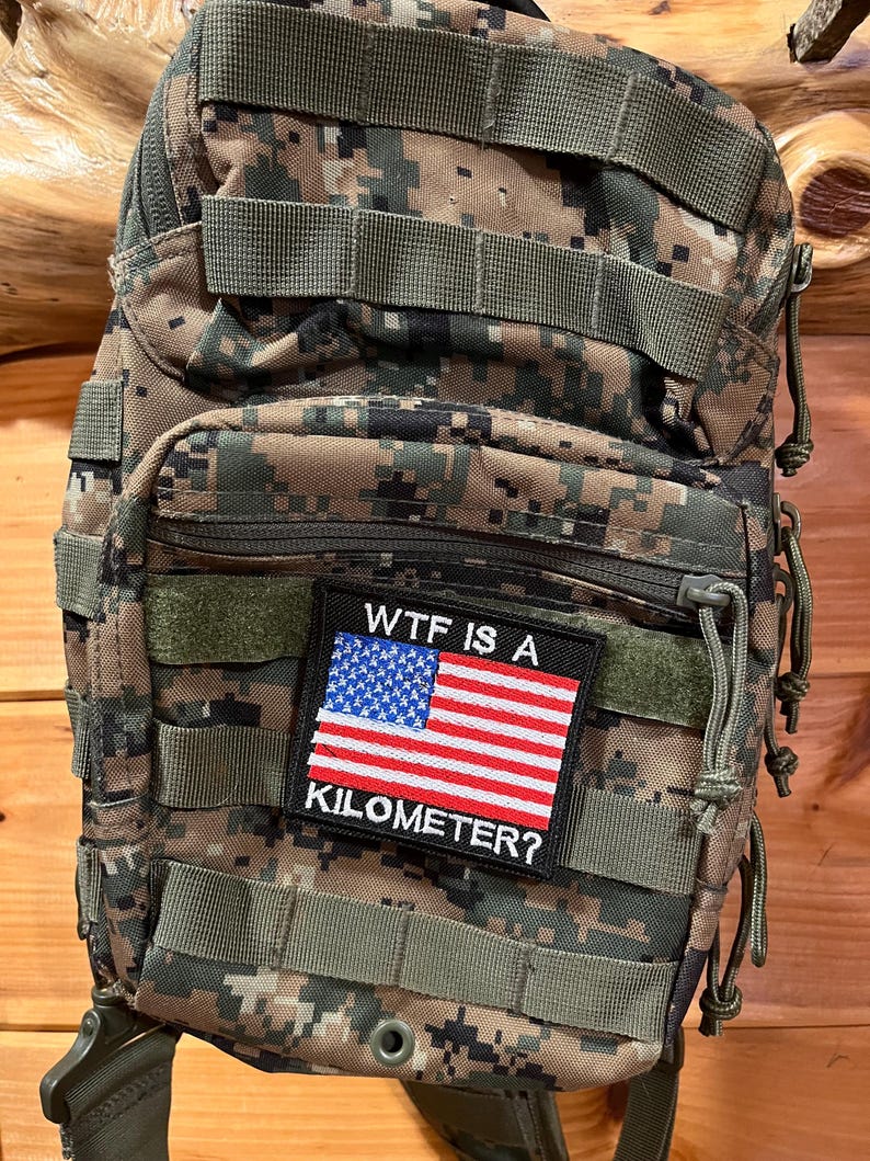 Pode incluir: Uma mochila t&aacute;tica de camuflagem com um patch que diz "WTF IS A KILOMETER?" e apresenta uma pequena bandeira americana.