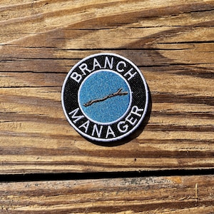 May include: Circular patch with the text "BRANCH MANAGER" in white on a black border. The centre features a blue background and a brown twig design. The patch is displayed on a wooden surface.