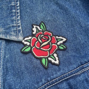 May include: A red embroidered rose patch with green leaves and white stitching on a blue denim jacket.