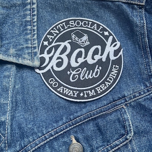Puede incluir: Un parche bordado en blanco y negro con el texto "Anti-Social Book Club Go Away I'm Reading" en una chaqueta vaquera azul.