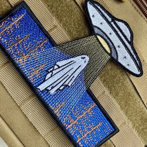 May include: A blue and brown embroidered patch with a white ghost being abducted by a grey UFO. The patch has the text "They're Coming" embroidered on it.