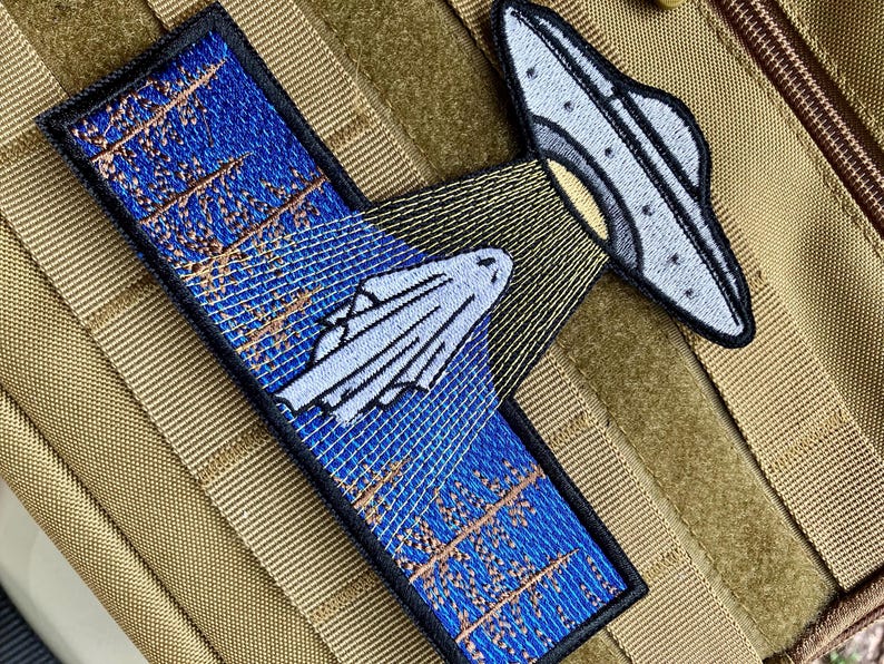 May include: A blue and brown embroidered patch with a white ghost being abducted by a grey UFO. The patch has the text "They're Coming" embroidered on it.