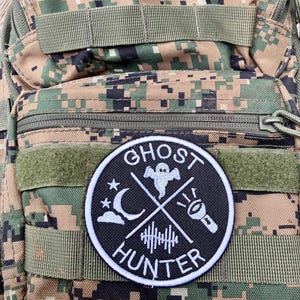 K&ouml;nnte beinhalten: Ein gestickter Patch in Schwarz und Wei&szlig; mit den Worten "Ghost Hunter" in einem Kreis. Der Patch zeigt ein Gespenst, einen zunehmenden Mond, eine Taschenlampe und Schallwellen.
