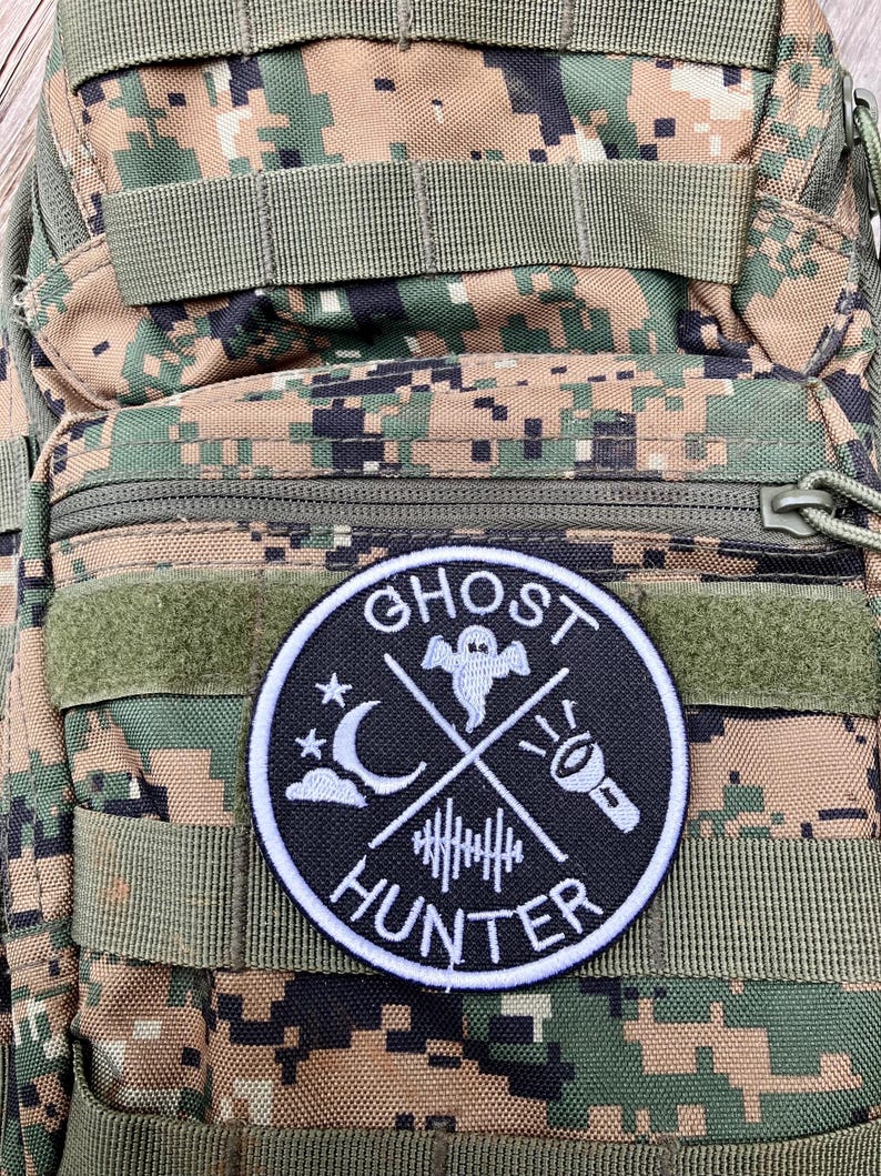 K&ouml;nnte beinhalten: Ein gestickter Patch in Schwarz und Wei&szlig; mit den Worten "Ghost Hunter" in einem Kreis. Der Patch zeigt ein Gespenst, einen zunehmenden Mond, eine Taschenlampe und Schallwellen.