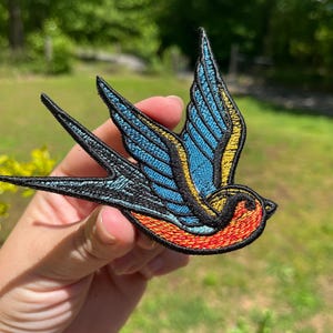 Puede incluir: Parche bordado con un diseño de golondrina en vuelo. El parche presenta detalles en azul, amarillo, naranja y negro. Las alas del ave están extendidas y el diseño está detallado con costuras.