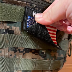 Pode incluir: Um remendo preto com suporte de velcro que apresenta uma pequena bandeira americana e a palavra "WAR" impressa nele.