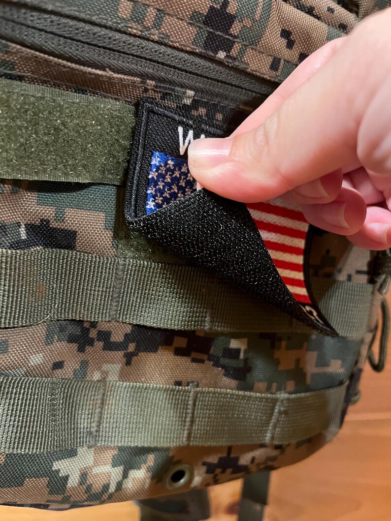 Pode incluir: Um remendo preto com suporte de velcro que apresenta uma pequena bandeira americana e a palavra "WAR" impressa nele.