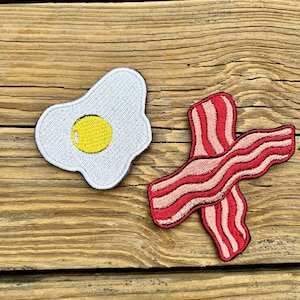 Può includere: Toppe ricamate raffiguranti un uovo fritto con tuorlo giallo e una striscia di pancetta con strisce rosse e rosa. Le toppe sono su una superficie di legno.