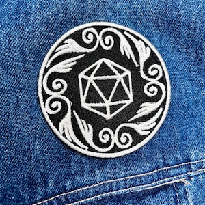 May include: A circular black patch with white embroidery. The design includes a central geometric shape, encircled by a white floral border. The patch is attached to a blue denim jacket.