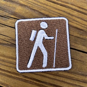 May include: A brown and white embroidered patch featuring a hiker with a backpack and walking stick. The patch is square with a white border, ideal for outdoor enthusiasts and those who enjoy hiking.