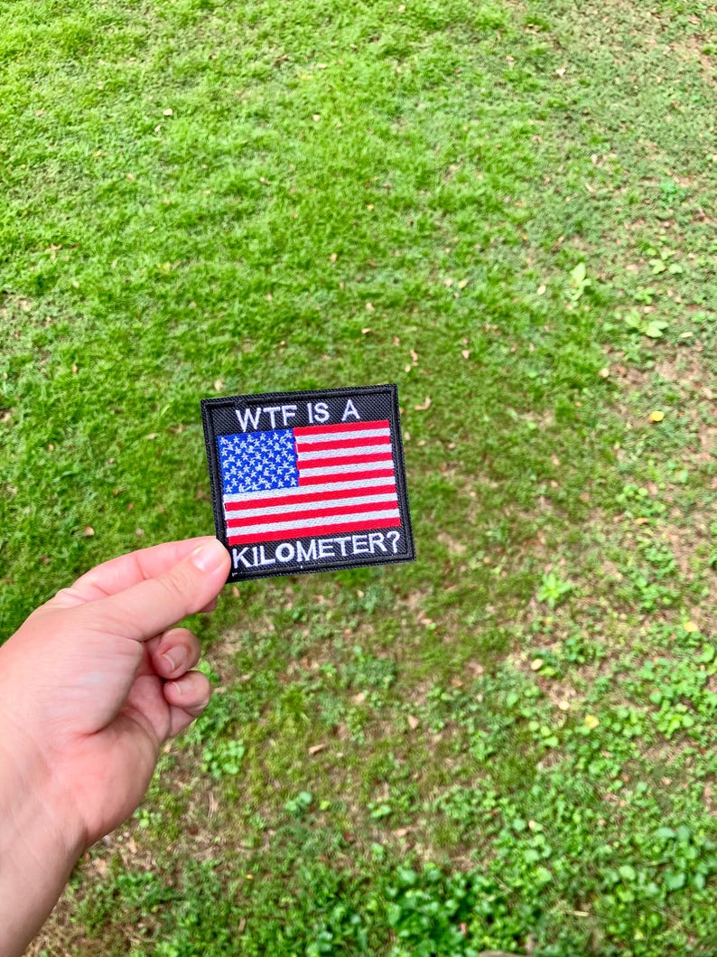 Pode incluir: Um remendo preto com a bandeira americana e o texto "WTF IS A KILOMETER?"