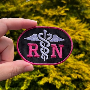 May include: An oval patch with a black background and a pink border. The patch features a white caduceus symbol and the letters "RN" in pink. The patch is held up against a blurred green background.