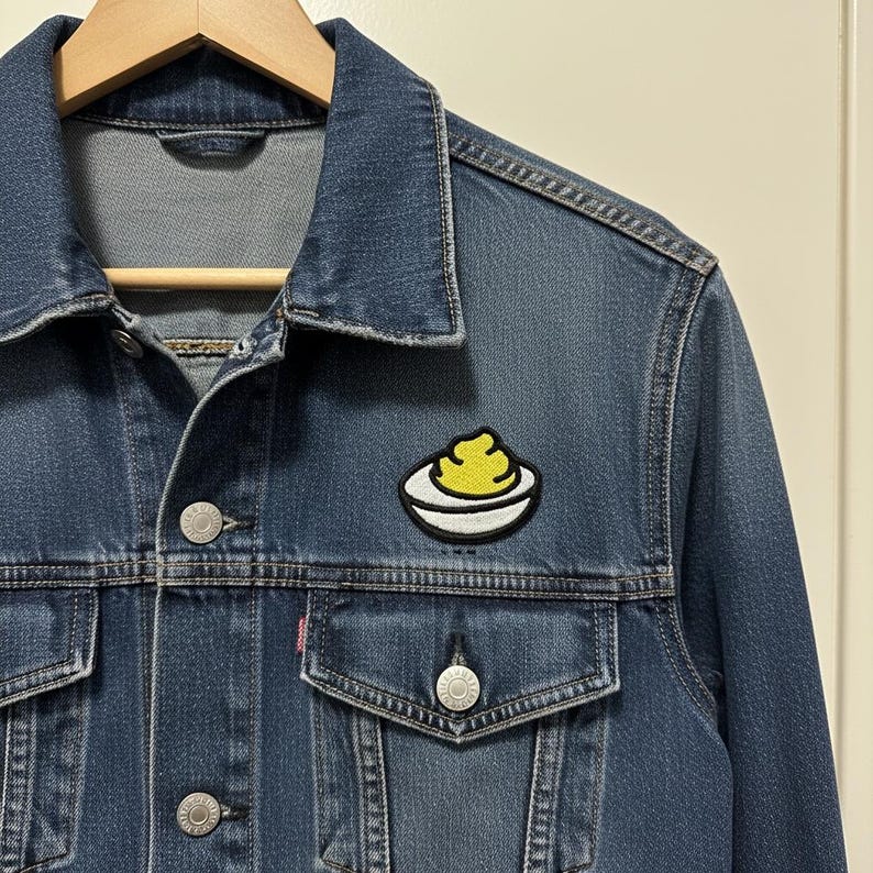 Puede incluir: Chaqueta vaquera azul medio con un peque&ntilde;o parche bordado. El parche presenta un alimento amarillo sobre un plato blanco con un contorno negro. La chaqueta tiene botones plateados y dos bolsillos en el pecho.
