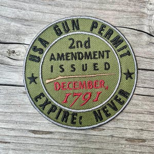 以下が含まれることがあります： 「USA GUN PERMIT 2nd AMENDMENT ISSUED」と黒字で「EXPIRE NEVER」と書かれた円形の緑色のパッチ。赤字で「DECEMBER, 1791」と書かれています。中央にはライフル銃のグラフィック。パッチには白い縁取りがあります。
