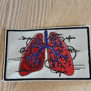 May include: A beige fabric patch with an embroidered anatomical illustration of a pair of lungs. The lungs are red with blue veins and are outlined in black. The patch has a black border and is stitched with white thread.