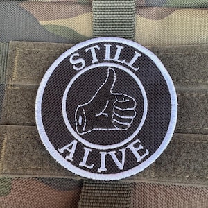 Peut inclure: Écusson rond noir et blanc avec les mots « STILL ALIVE » et un pouce levé. L'écusson est cousu sur un tissu de camouflage.