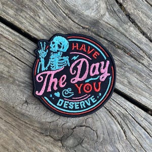 May include: A black and white embroidered patch featuring a skeleton holding up a peace sign. The patch reads "Have the Day You Deserve" in pink and red lettering.