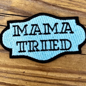 May include: A light blue embroidered patch with the words "MAMA TRIED" in black block letters. The patch has a black border and a decorative shape. The patch is on a wooden surface.