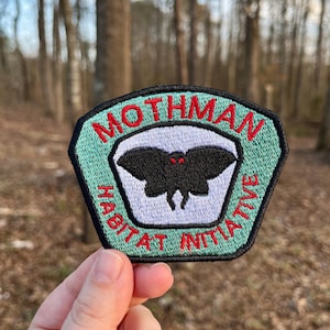 Può includere: Una toppa esagonale verde con le parole "Mothman Habitat Initiative" ricamate in rosso attorno al bordo. Una falena nera con occhi rossi è ricamata al centro della toppa.