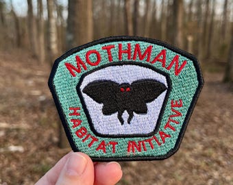 Puede incluir: Un parche hexagonal verde con las palabras "Mothman Habitat Initiative" bordadas en rojo alrededor del borde. Una polilla negra con ojos rojos está bordada en el centro del parche.