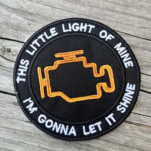 Puede incluir: Parche redondo negro con texto blanco bordado: "THIS LITTLE LIGHT OF MINE I'M GONNA LET IT SHINE". El parche presenta un símbolo de motor bordado en naranja en el centro.