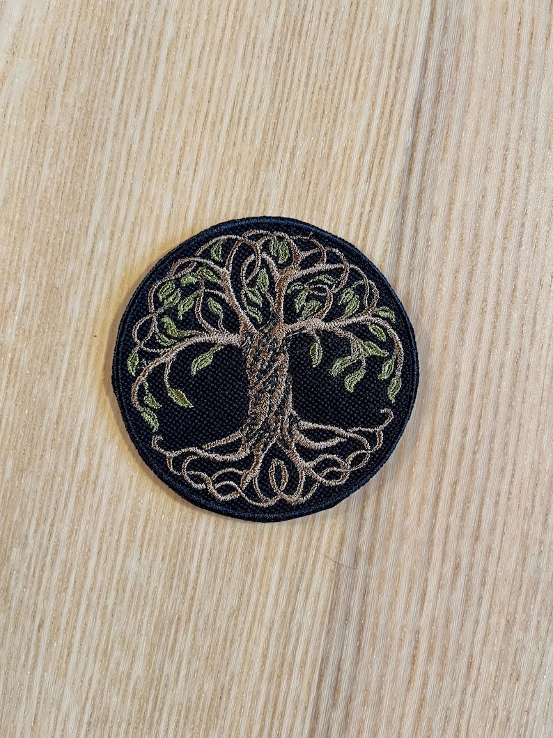 May include: Circular embroidered patch with a tree of life motif. The tree is depicted with a brown trunk and green leaves, set on a dark blue circular background. Ideal for embellishing clothing or accessories.