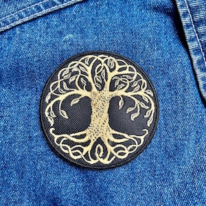 May include: A round, black embroidered patch featuring a gold-colored tree design. The tree has a detailed trunk, branches, and leaves. The patch is set against a blue denim background.