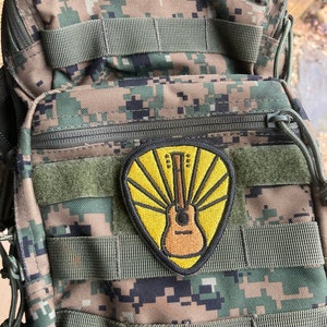 Może przedstawiać: Kamuflażowy plecak z ż&oacute;łto-brązową naszywką gitary. Naszywka ma wz&oacute;r promieni słonecznych z gitarą w środku.