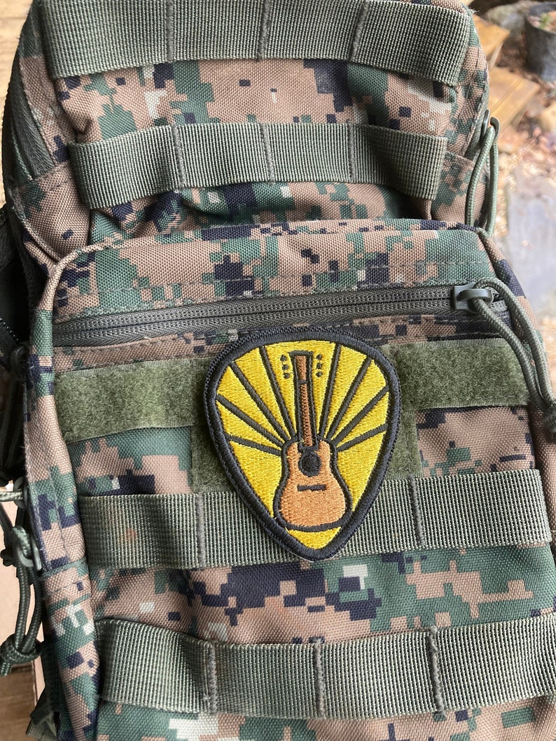 Może przedstawiać: Kamuflażowy plecak z ż&oacute;łto-brązową naszywką gitary. Naszywka ma wz&oacute;r promieni słonecznych z gitarą w środku.